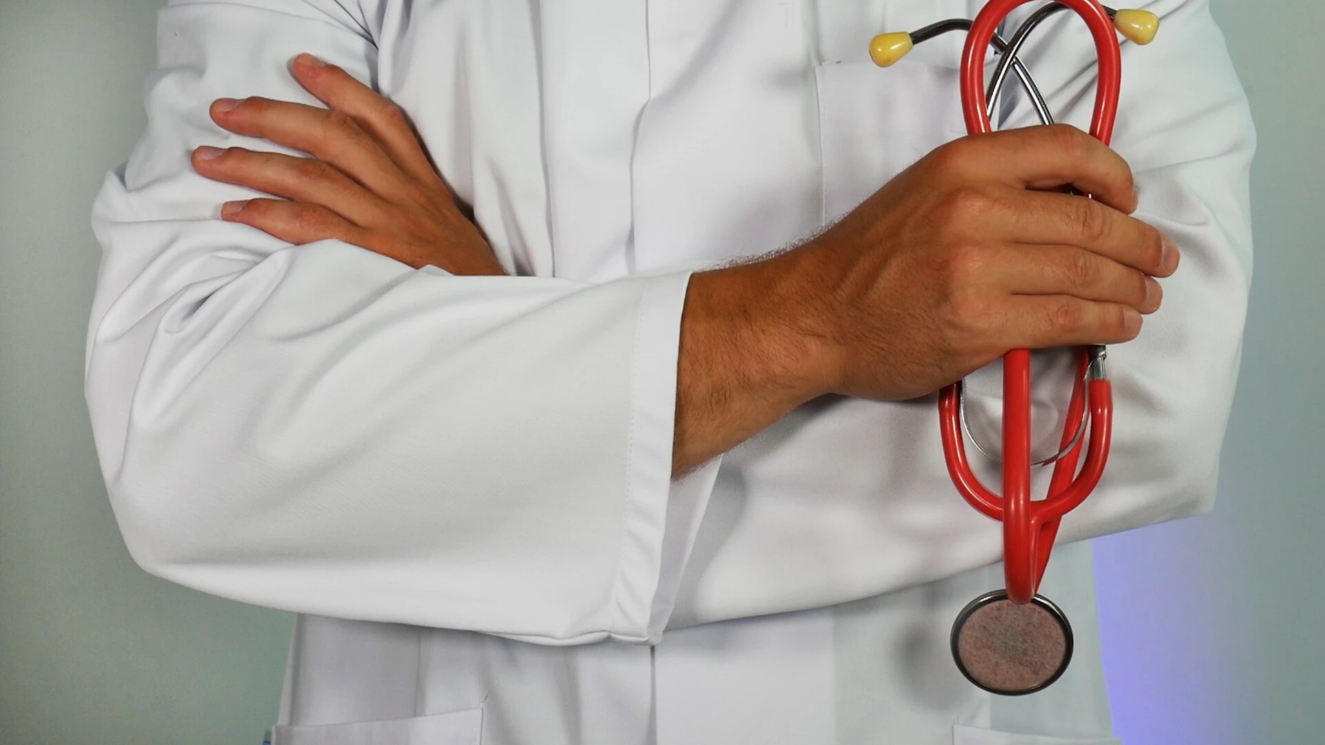 A doctor in a white lab coat stands with crossed arms, holding a red stethoscope.