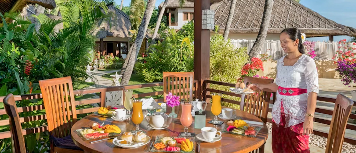 A smiling woman serves a vibrant tropical breakfast at an outdoor table in a Bali resort.