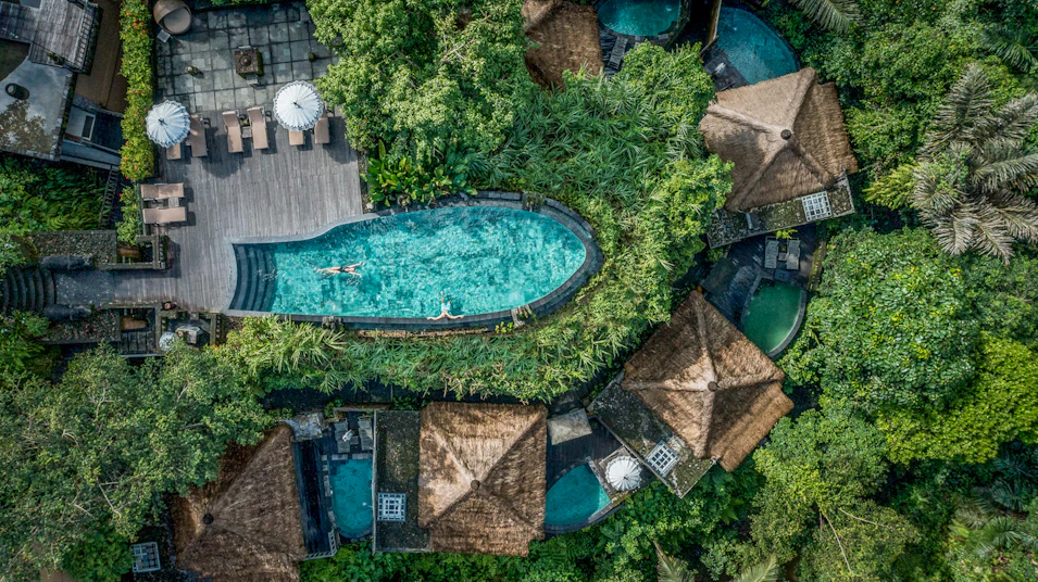 Two people swim in a stunning resort pool surrounded by lush jungle and traditional bungalows in Bali.