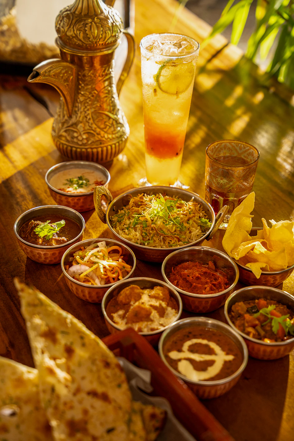 A lavish array of Indian curries, rice, naan bread, and a colorful drink on a wooden table.