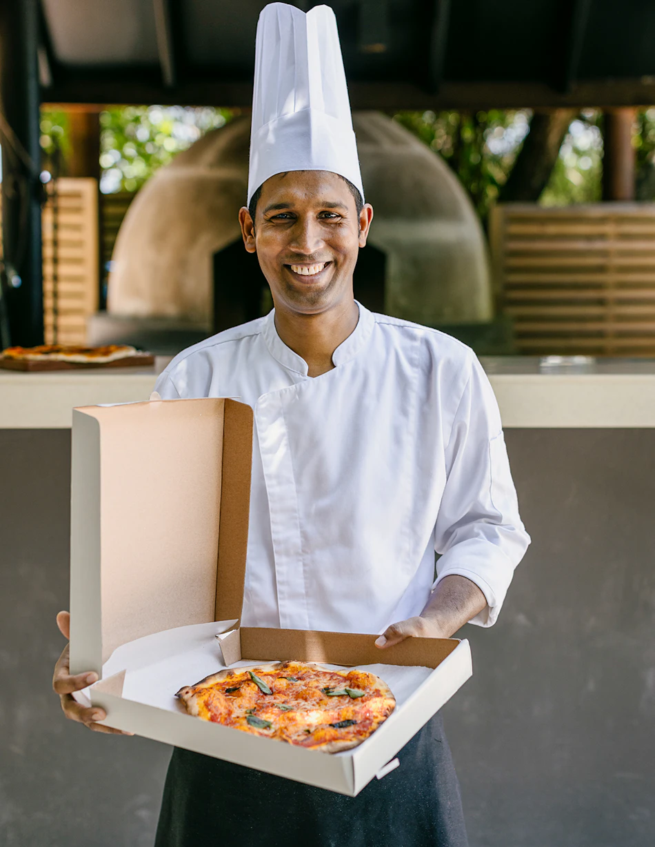 A smiling resort chef holds a freshly baked pizza in an open box near a wood-fired oven.