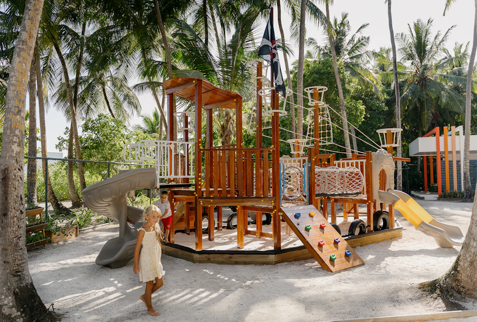 Children enjoy a large pirate ship playground with slides and nets on a sandy tropical resort.