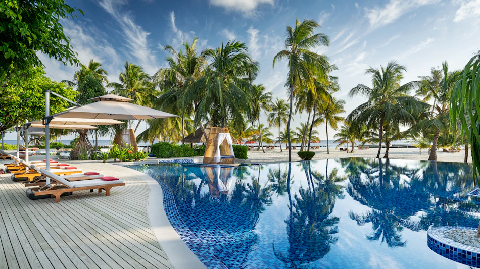 Serene resort swimming pool with lounge chairs and palm trees leading to a sandy Maldives beach.