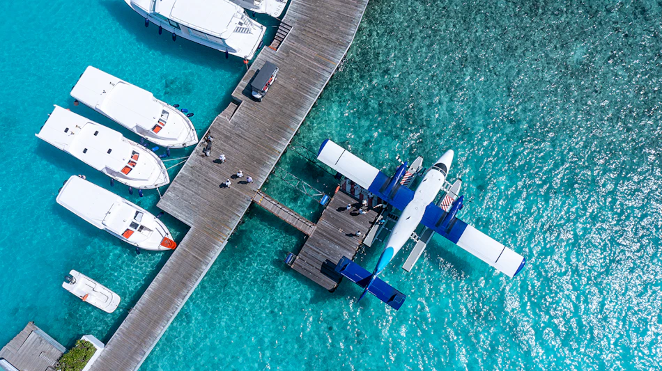 Seaplane and white yachts docked at a long wooden pier over clear turquoise waters in the Maldives.