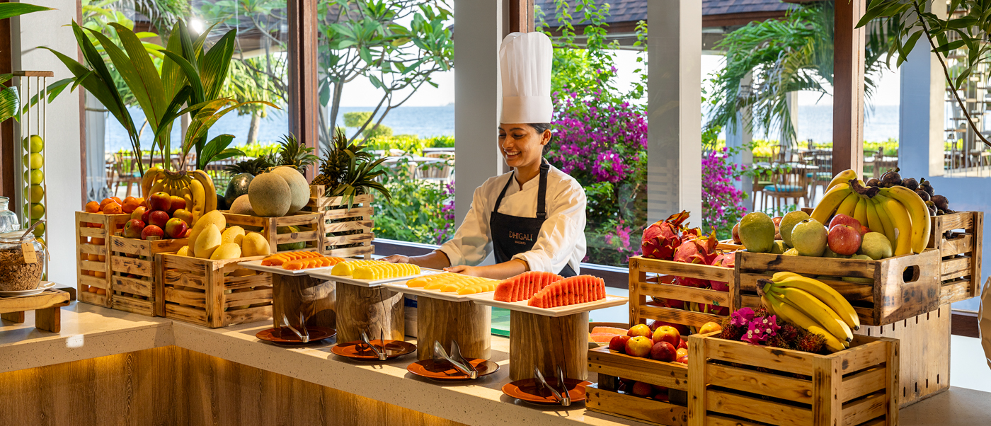 A smiling chef stands behind a vibrant tropical fruit buffet with an ocean view in the Maldives.