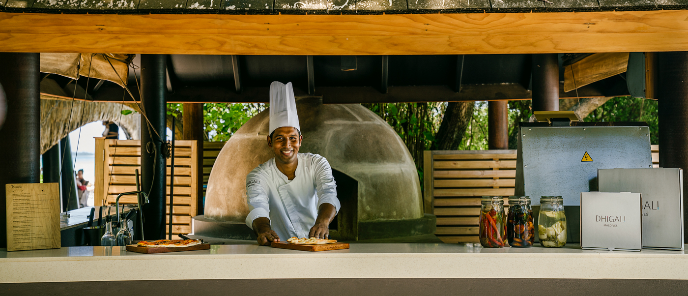 A smiling chef prepares pizza in an outdoor kitchen with a large oven at Dhigali Maldives.