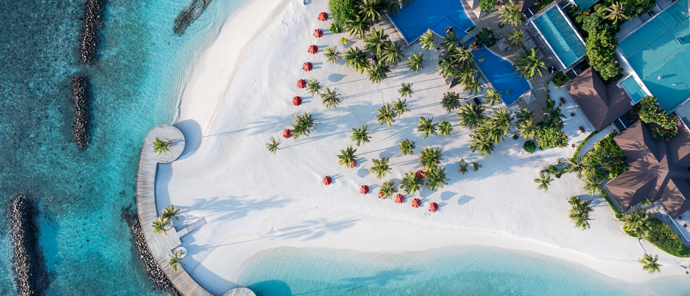 Aerial view of Dhigali Maldives resort with white sand beach, palm trees, red umbrellas, and turquoise water.