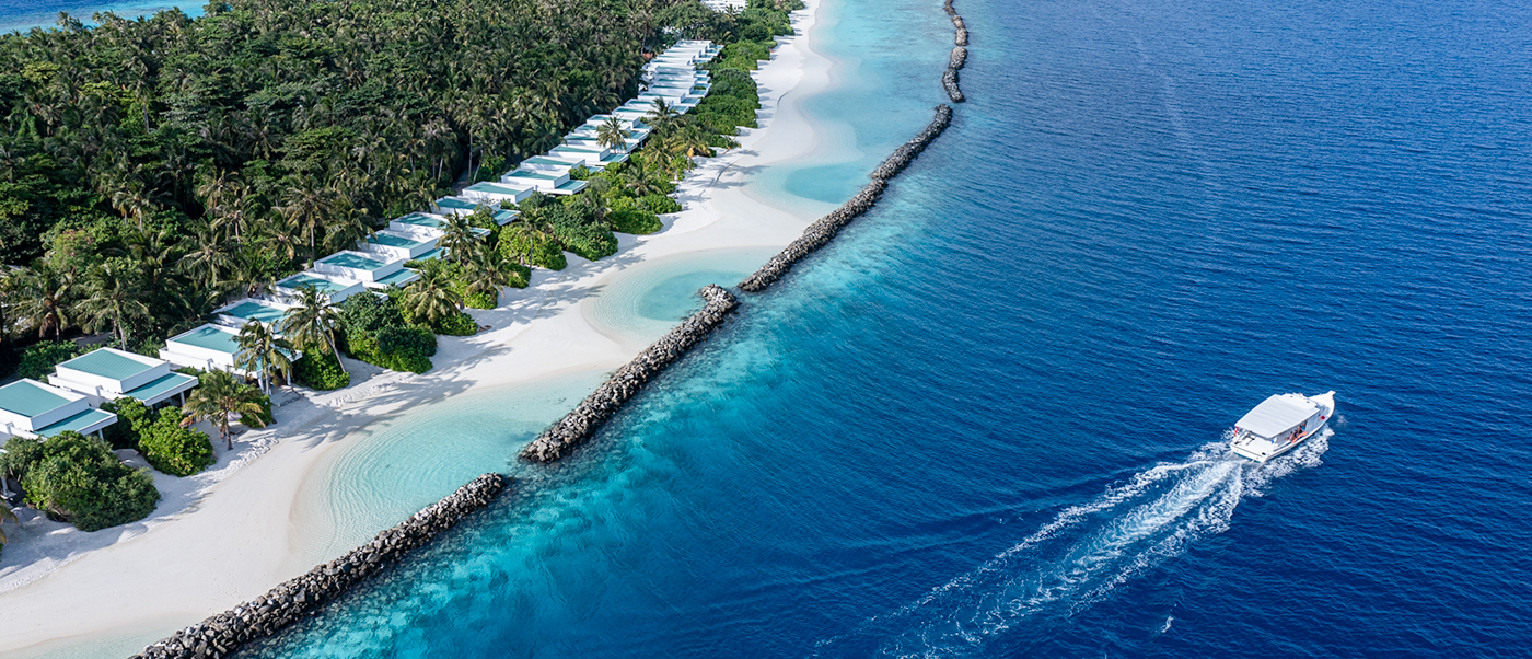 Aerial view of Dhigali Maldives resort, featuring white villas, sandy beaches, and a boat on the turquoise ocean.