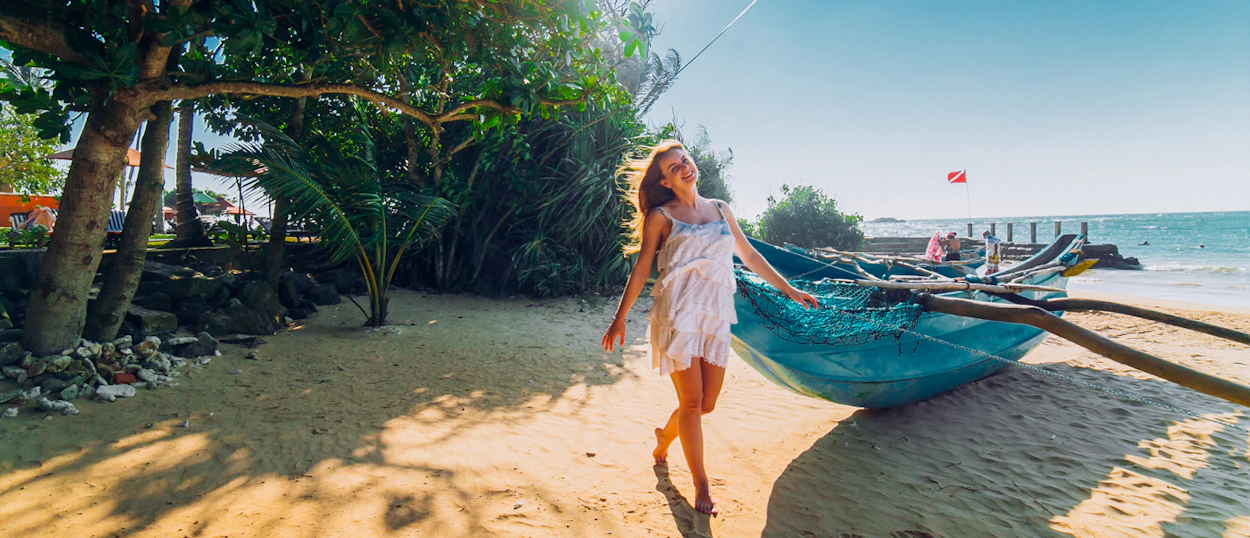 A smiling woman walks on a sunny golden beach with a blue fishing boat in Weligama.