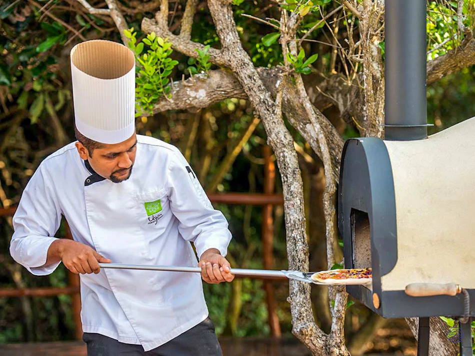 A chef carefully places a pizza into a wood-fired oven outdoors at Uga Chena Huts.