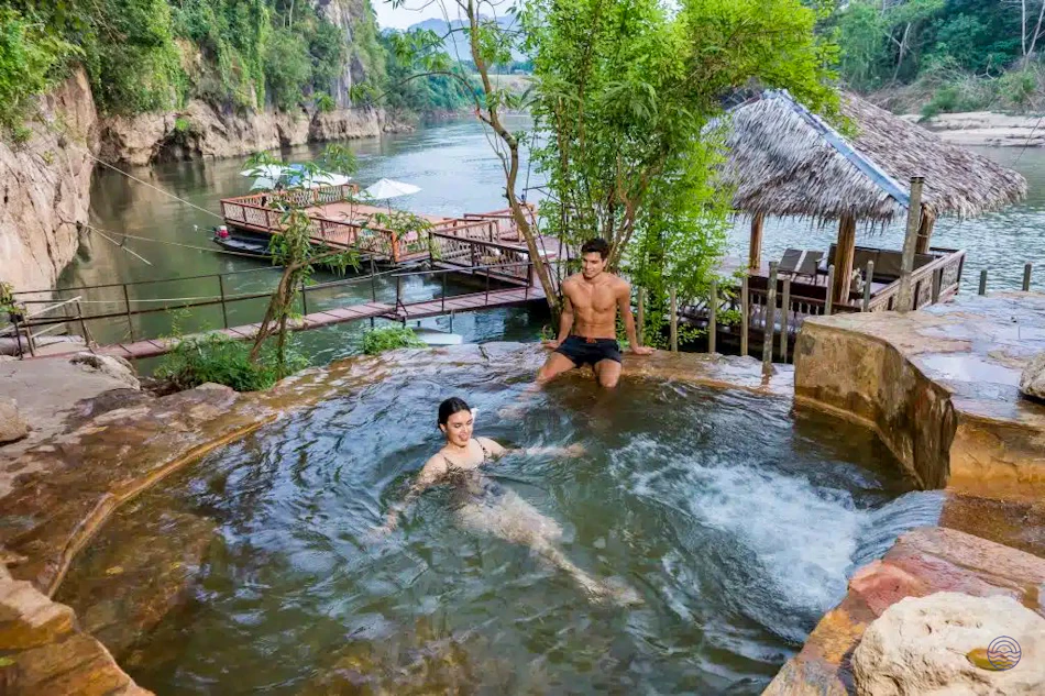 A couple relaxes in a natural hot spring pool overlooking a serene river in Kanchanaburi, Thailand.