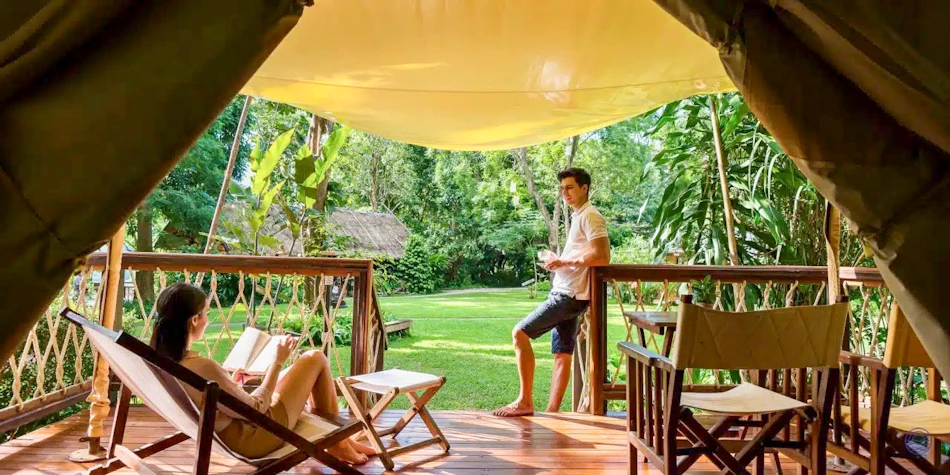 A couple relaxes on a tented lodge deck, enjoying a book and the lush tropical view.