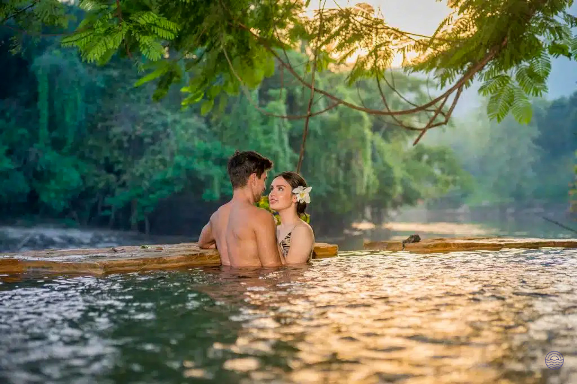 A couple shares a tender moment, submerged in a sunlit river with lush green trees.