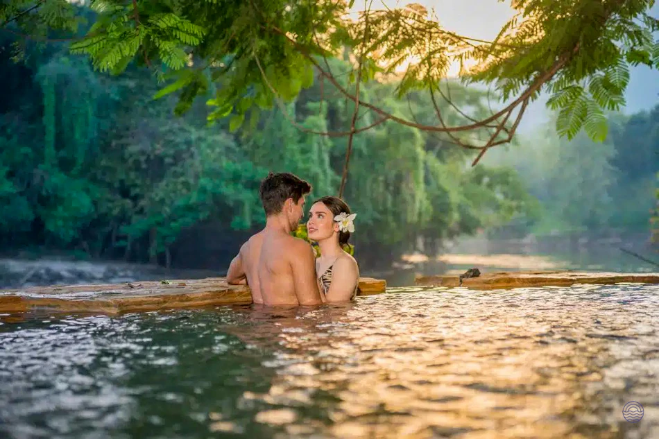 A couple shares a tender moment, submerged in a sunlit river with lush green trees.
