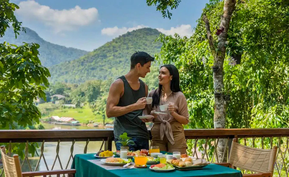A happy couple enjoys breakfast on a balcony overlooking a scenic river and lush mountains.