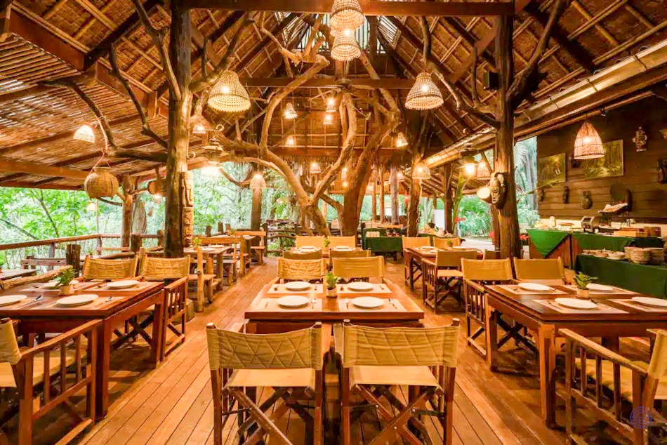 Rustic open-air restaurant at Hintok River Camp with wooden tables, thatched roof, and integrated trees.