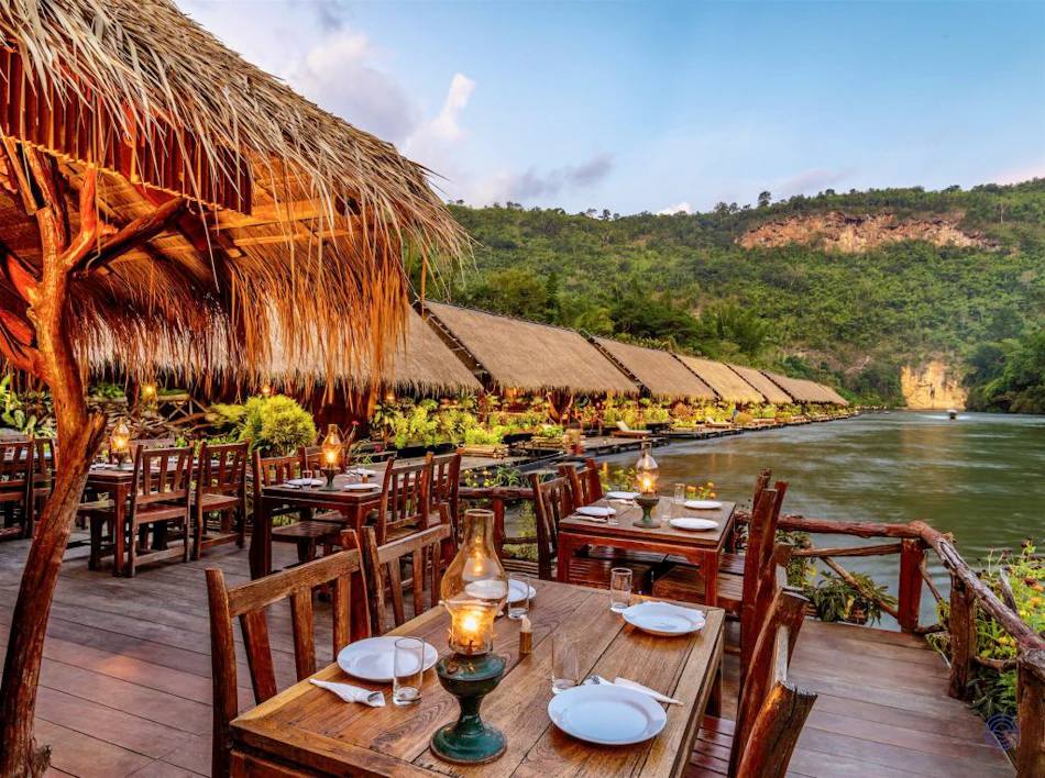 Romantic thatched-roof restaurant with wooden tables and lanterns overlooking the scenic River Kwai and jungle hills.