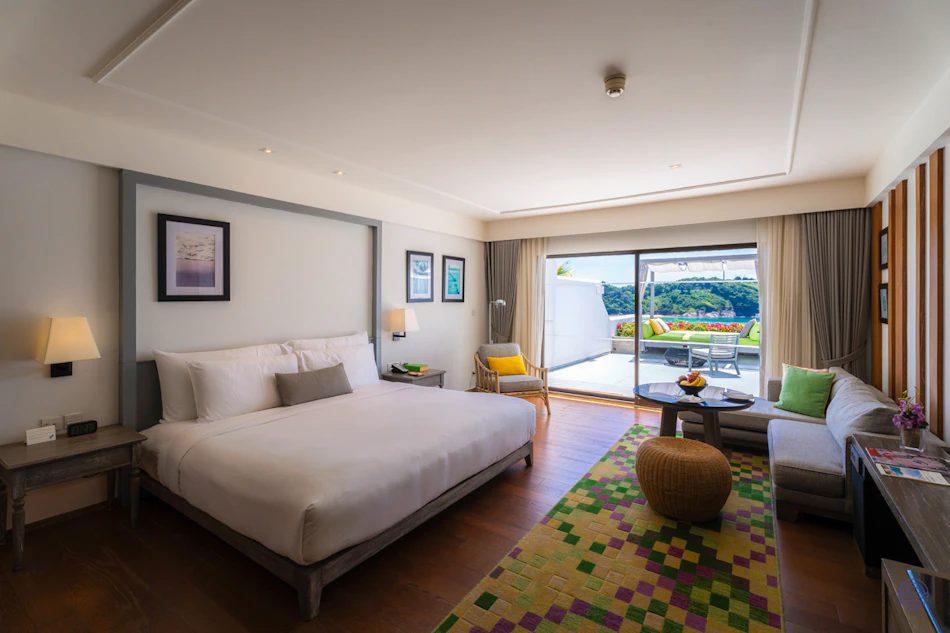 Luxurious hotel room with a king bed, lounge area, and a balcony view of Phuket's green hills.