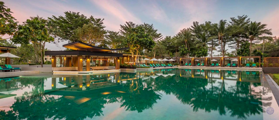 Luxurious resort swimming pool and bar area surrounded by tropical trees at sunset in Thailand.