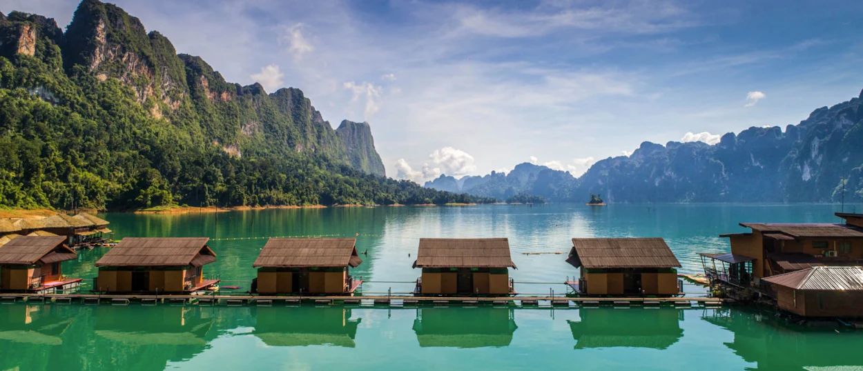Serene floating bungalows on Chiewlan Lake, Khao Sok, reflecting towering limestone karsts under a bright blue sky.