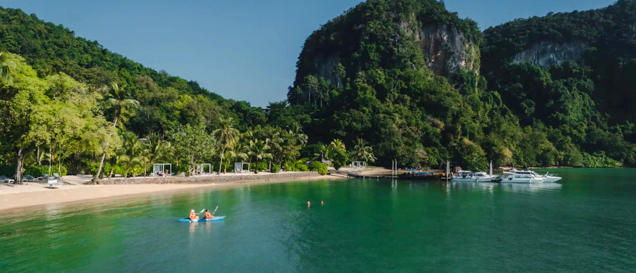 Kayakers paddle on calm turquoise water off a serene Koh Yao Noi beach with lush green cliffs.