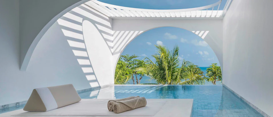 A modern white private pool villa overlooking the ocean with tropical palm trees and blue sky.