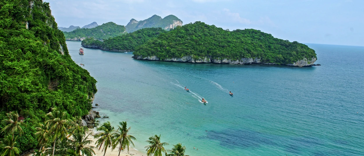 Boats cruise past lush green islands and a sandy beach with palm trees in Koh Samui.