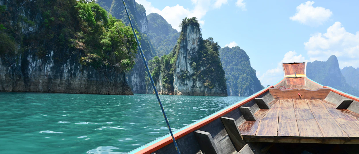 A longtail boat glides on turquoise water past towering limestone karsts under a bright sky.