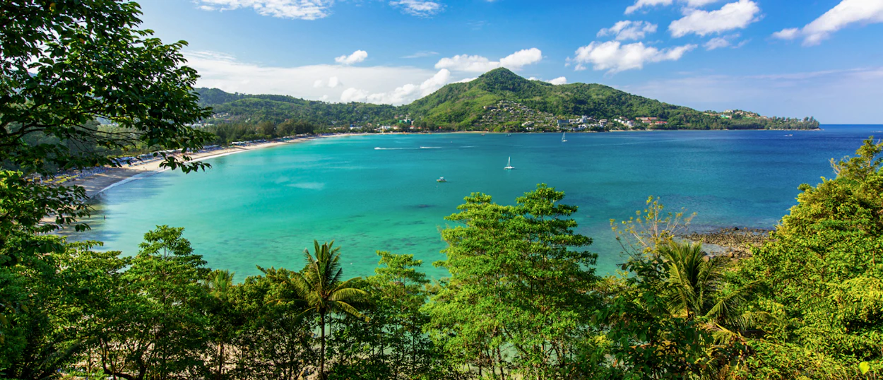 A serene tropical bay in Phuket with clear turquoise water, a sandy beach, and green hills.