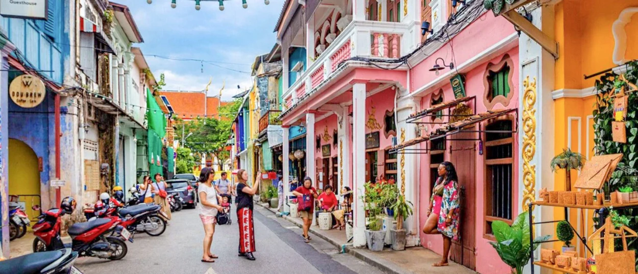 Tourists explore a vibrant street in Phuket Old Town with colorful Sino-Portuguese buildings and motorbikes.