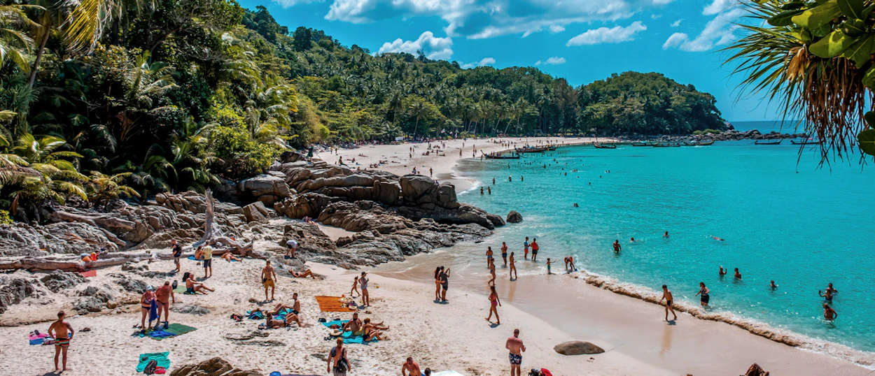 People relax and swim on a sunny tropical beach with turquoise water and green hills in Phuket.