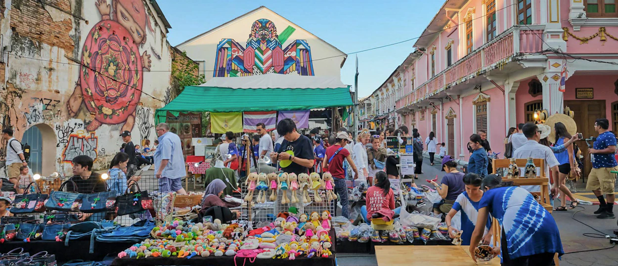 A bustling street market in Phuket Old Town with people browsing colorful stalls and unique street art.