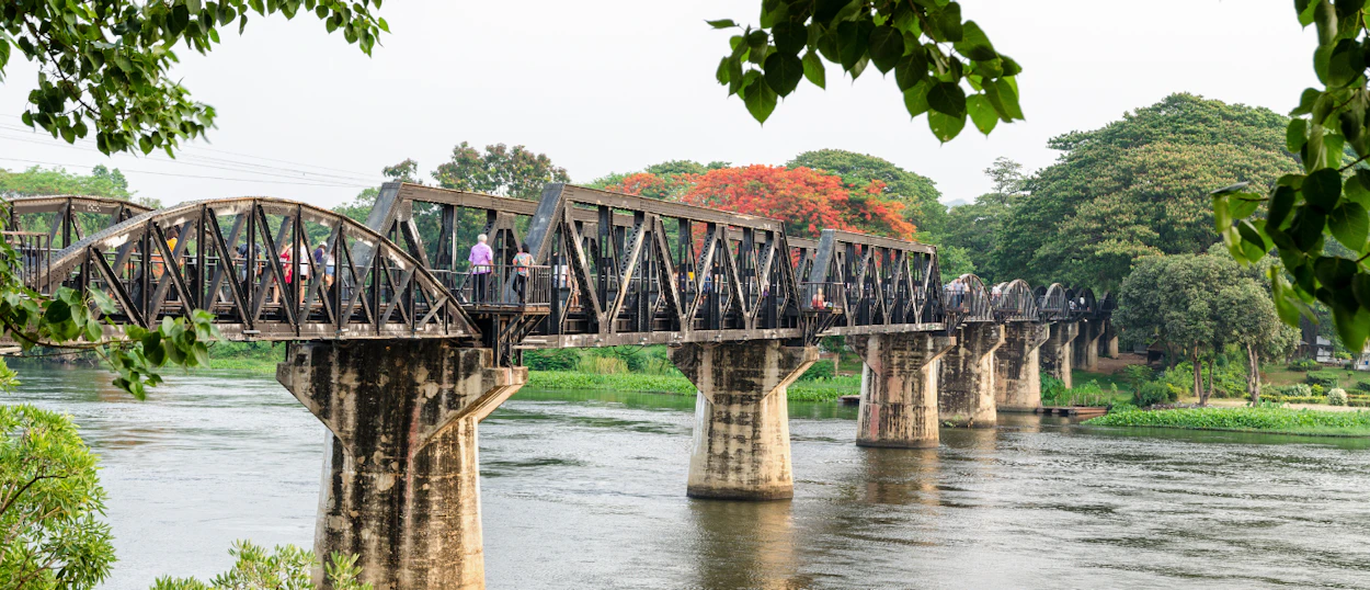 Tourists walk on the historic Bridge on the River Kwai in Kanchanaburi, Thailand, framed by lush trees.