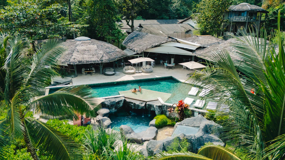 Lush tropical resort in Thailand featuring a multi-level pool, waterfall, and thatched-roof bungalows.