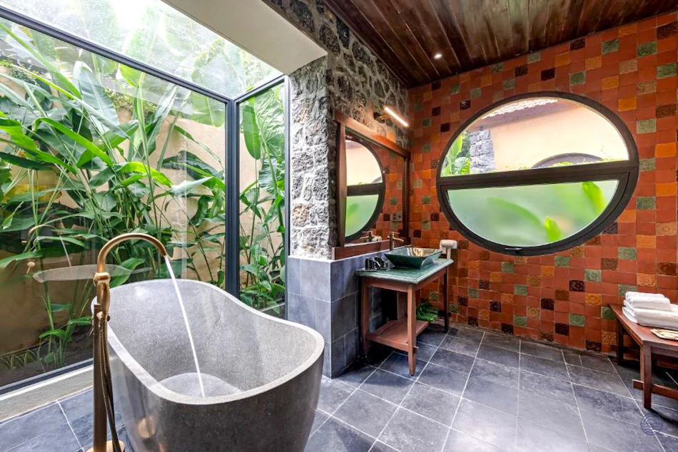 Luxurious bathroom with a stone tub, running water, and a glass wall overlooking tropical plants.