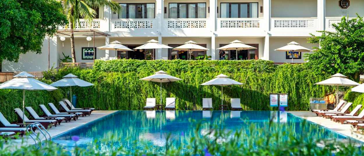 A peaceful resort swimming pool in Hoi An with sun loungers, umbrellas, and lush green foliage.