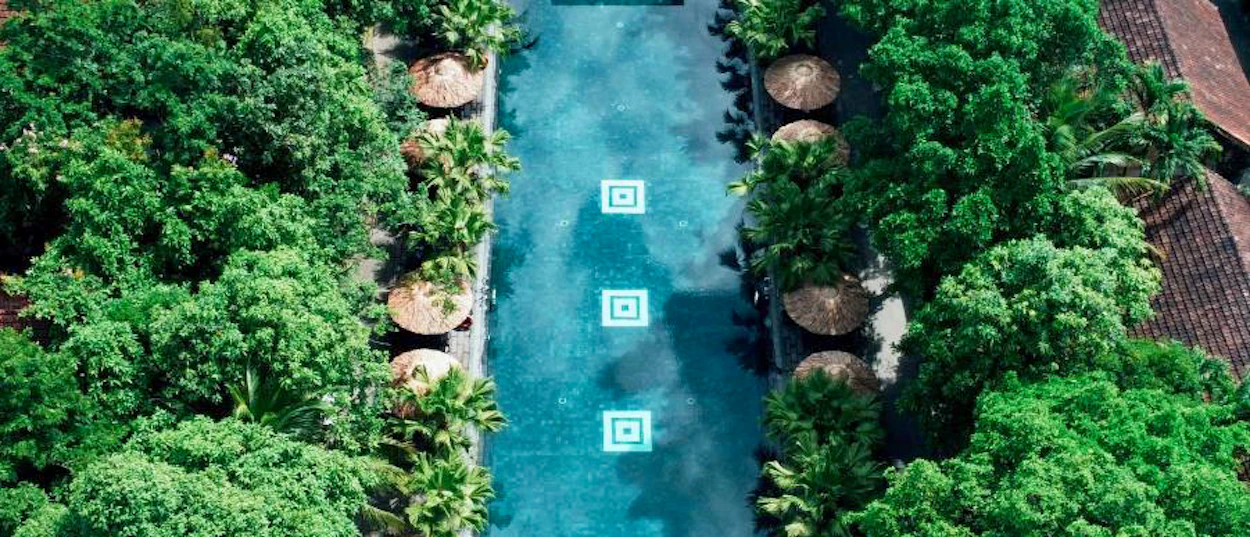 Aerial view of a long resort swimming pool surrounded by lush green trees and cabanas in Hue.