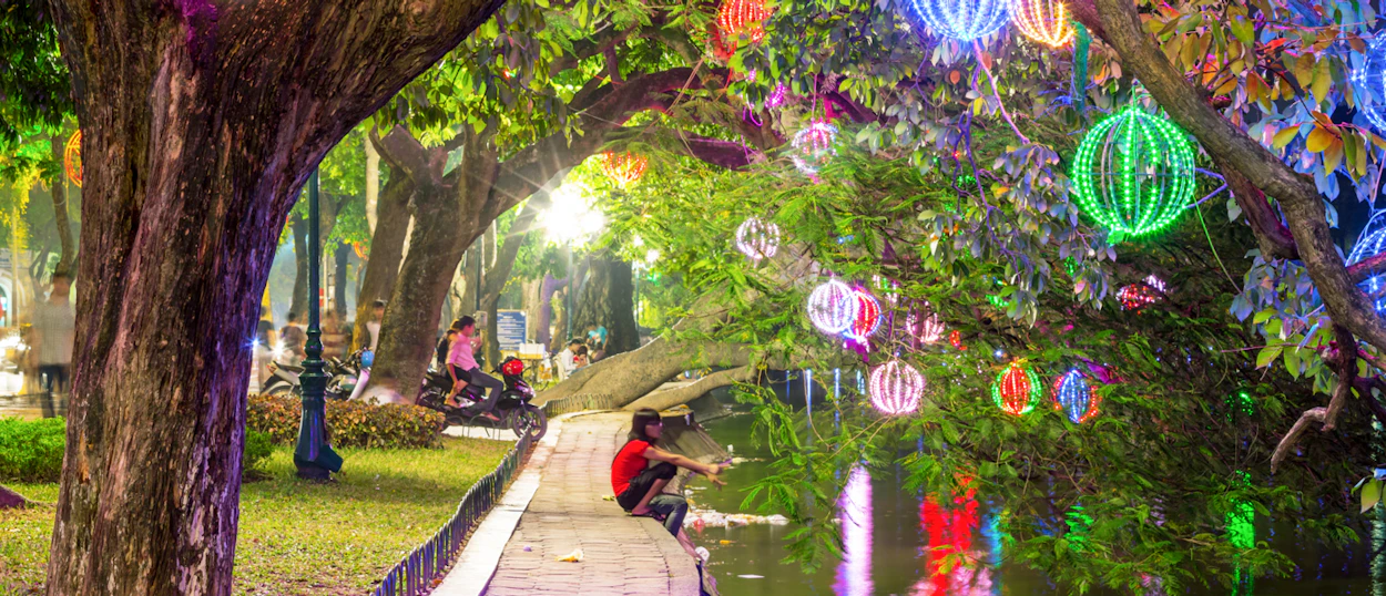 Colorful glowing lanterns adorn trees and reflect in water, creating a festive evening scene in Hanoi.