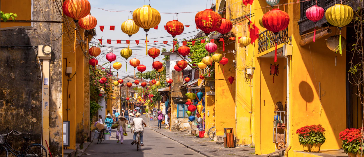 Colorful lanterns hang above a bustling street in Hoi An, lined with historic yellow buildings.