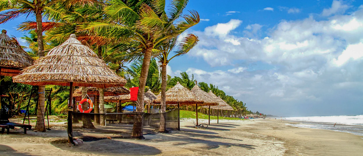 Tropical Thuan An Beach with palm trees, straw umbrellas, and gentle waves under a bright sky.