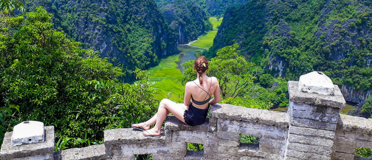A woman sits on a stone wall, admiring the panoramic view of Tam Coc's green valley and karsts.